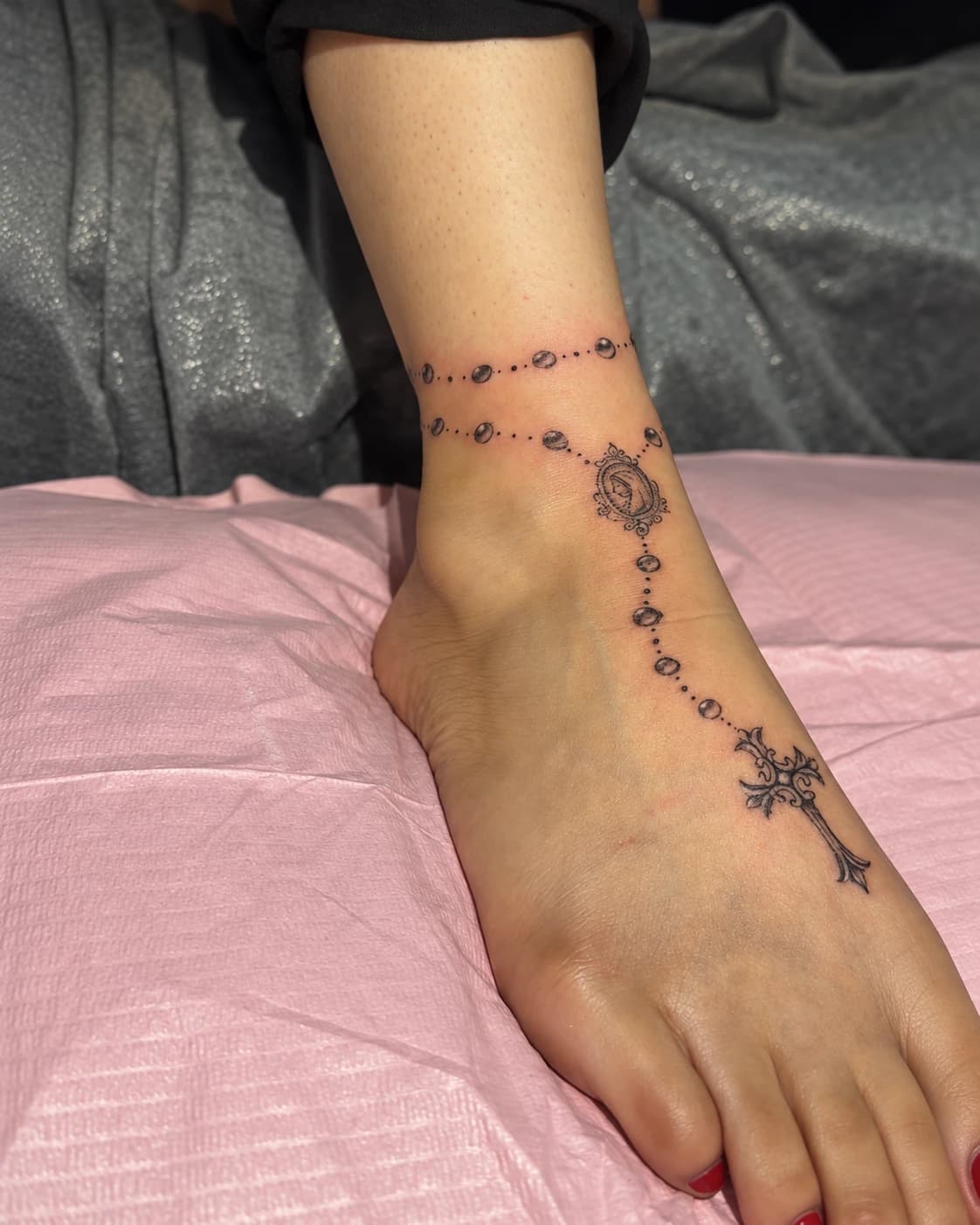 A rosary tattoo circling an ankle with a cross extending onto the foot